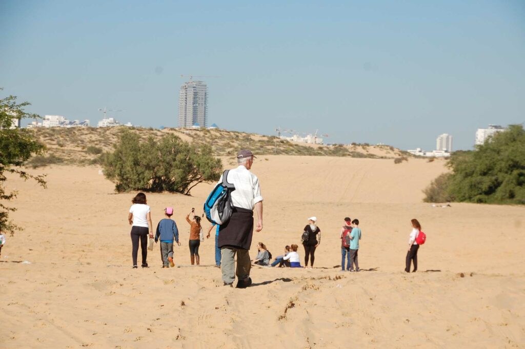 מטיילים וילדים בדיונה באשדוד על רקע הבניינים של העיר 