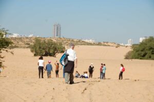 מטיילים וילדים בדיונה באשדוד על רקע הבניינים של העיר